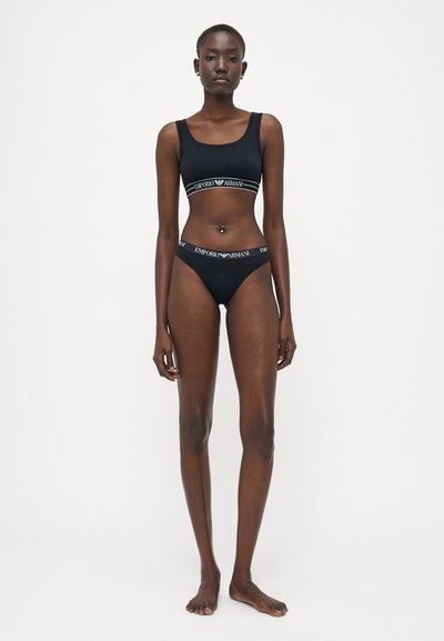 Black sports bra and matching thong with "Emporio Armani" logo in white. Smooth texture, fitted design, wide straps, low-rise cut.