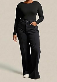 Personne portant un haut noir à manches longues et un pantalon noir évasé taille haute avec des chaussures blanches, debout avec une main dans la poche.
