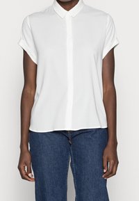 Une personne à la peau foncée portant une chemise blanche à manches courtes avec des poignets retroussés et un jean en denim bleu, se tenant devant un fond uni.