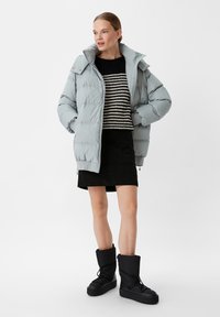 Hellgraue Daunenjacke mit hohem Kragen und Reißverschluss, kombiniert mit einem schwarz-cremefarben gestreiften Strickpullover und einem schwarzen, strukturierten Rock. Schwarze Stiefeletten.