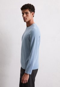 Helles blaues Langarmshirt aus weichem Stoff mit Rundhalsausschnitt. Figurschnitt und dezente Nähte, kombiniert mit dunklen Hosen.