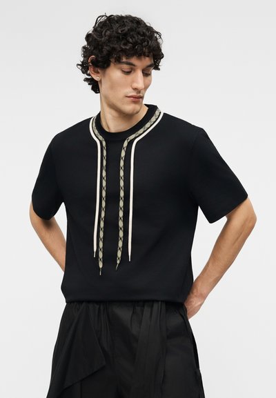 Νεαρός άνδρας με σγουρά μαλλιά φοράει μαύρο πουκάμισο με κοντά μανίκια, μακριά κορδόνια με σχέδια και μαύρο φαρδύ παντελόνι, κοιτάζοντας προς τα κάτω.