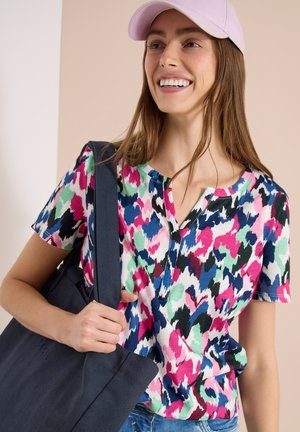 Femme souriante portant une casquette rose, une chemise à manches courtes à motifs colorés, tenant un grand sac cabas foncé sur une épaule.