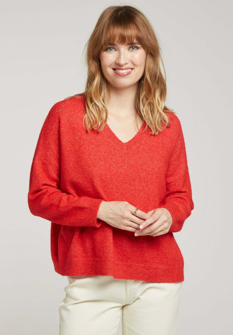 Pull en v rouge surdimensionné avec de longues manches, tissu texturé, poignets et ourlet côtelés. Porté sur un pantalon beige clair.