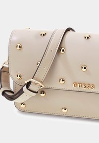 Sac à main beige avec clous dorés, doté d'une fermeture à rabat et d'une bandoulière réglable. Nom de la marque "GUESS" affiché en doré sur le devant.