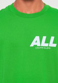 Artimiausias planas, kuriame matomas asmuo, vilkintis žalią marškinėlį su balta užrašu "ALL" ir "CALVIN KLEIN", atspaustu ant krūtinės.