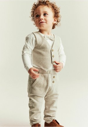 Toddler with curly hair wearing beige vest, matching pants with buttons, white long-sleeve shirt, bow tie, and brown shoes, standing indoors.
