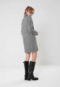 Gray, knitted sweater dress with a high collar, knee-length. Paired with shiny black knee-high boots featuring a buckle detail.