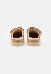 Bež zamšaste cipele na vezanje s mekim unutarnjim oblogom, petnom petljom i svijetložutim gumenim potplatima. UGG logo straga.