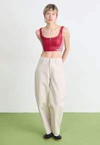 Top curto vermelho com fecho e riscas brancas, combinado com calças largas bege e sapatilhas pretas. Modelo está em pé sobre um pavimento verde contra um fundo branco.