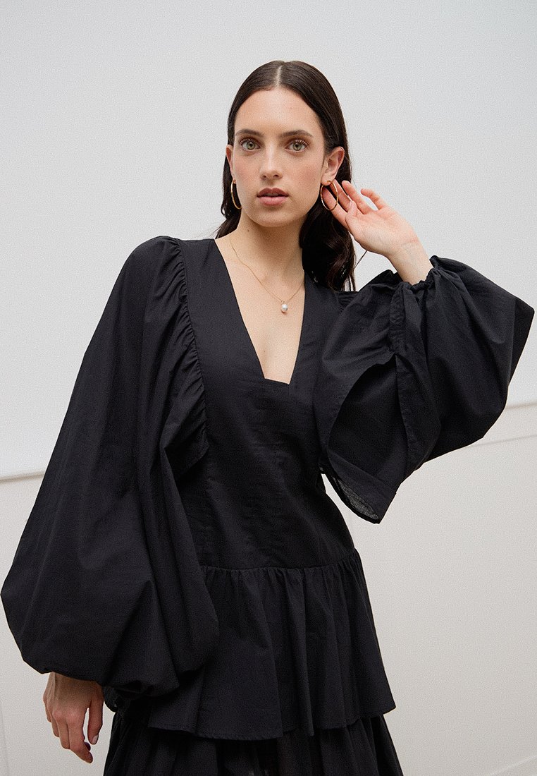 Schwarzes Peplum-Oberteil mit tiefem V-Ausschnitt, gepufften langen Ärmeln und gerafften Details, hergestellt aus einem glatten Stoff mit strukturiertem Finish.