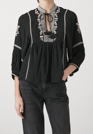 Blouse noire avec broderie florale blanche, manches trois-quarts plissées et détail de nœud à l'avant, portée avec un jean gris foncé.