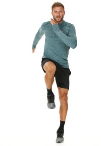 Atleet in een teal langemouwtop en zwarte shorts, halverwege de sprint, met zwarte sokken en grijze hardloopschoenen met gele accenten.