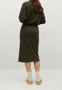 Kvinde med langt brunt hår iført en mørkegrøn langærmet kjole og cremefarvede ankelstøvler, der står foran en ensfarvet baggrund.