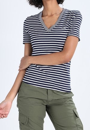 Femme portant un t-shirt à manches courtes rayé bleu marine et blanc avec un col en V et un logo, ainsi qu'un pantalon cargo vert olive, les bras croisés sur le torse, sans détails d'arrière-plan.