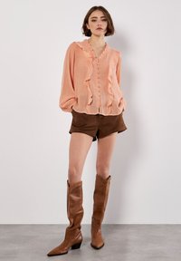 Blusa de melocotón con detalles de volantes y cierre de botón, combinada con pantalones cortos de ante marrón y botas de cuero marrón hasta la rodilla.