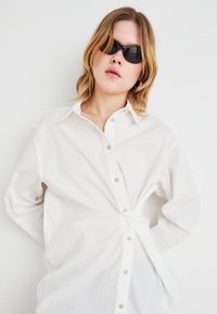 White button-up shirt with long sleeves and a tied front detail. Worn with black sunglasses. Smooth fabric texture, classic collar design.
