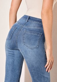 Zilas džinsa bikses ar pieguļošu dizainu, vidējā augstumā, divām aizmugurējām kabatām un smalkām svītrojumu detaļām. Gluda tekstūra un viegla izskata.