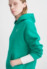 Zielona bluza z kapturem z kangurzą kieszenią, ściągaczami na rękawach oraz haftowanym logo na klatce piersiowej. Materiał wydaje się miękki i fakturowany.