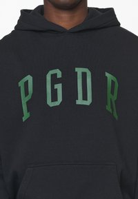Persona che indossa un felpa nera con grandi lettere verdi "PGDR" stampate sul petto.