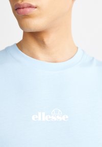 Gros plan sur une personne portant un t-shirt bleu clair avec un logo blanc "ellesse" centré sur la poitrine.