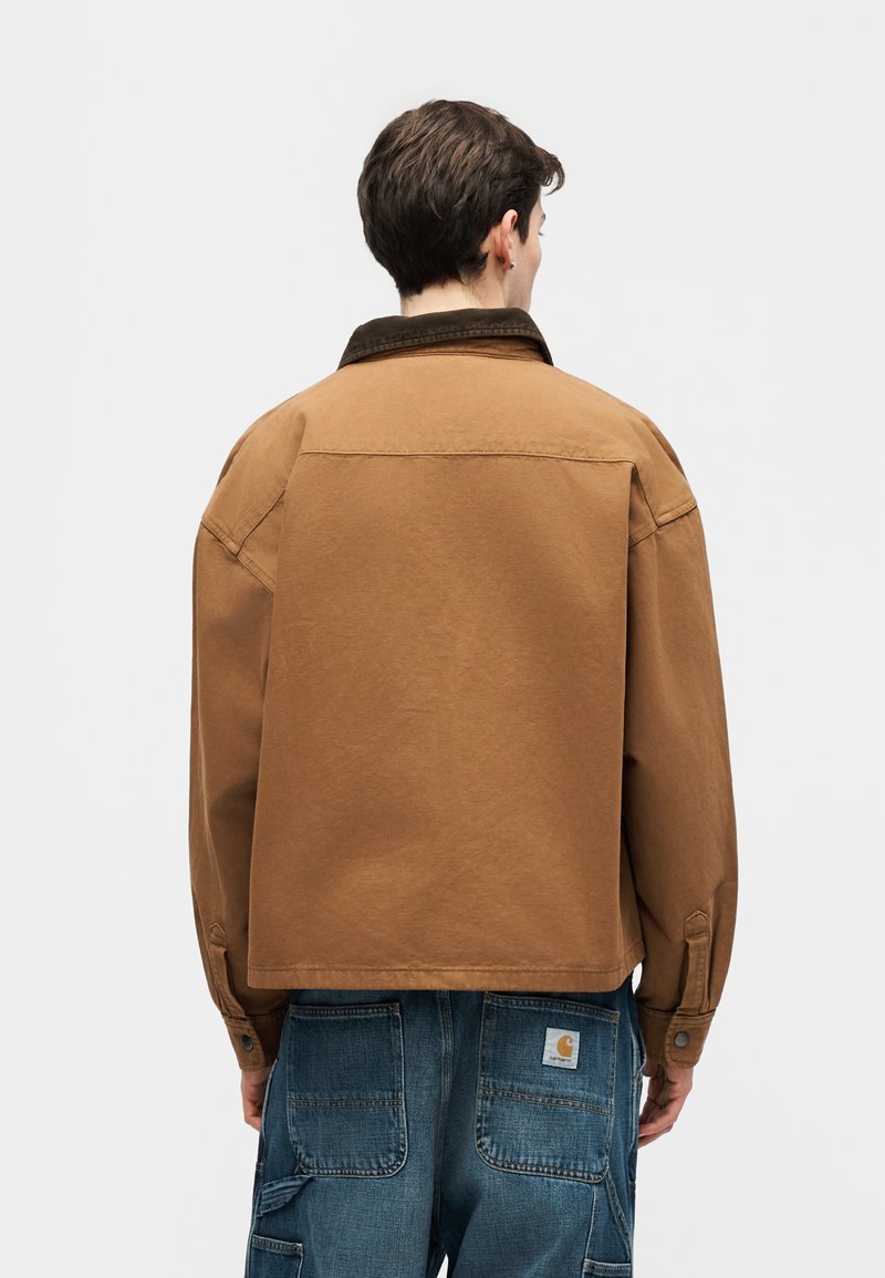 Person mit kurzen dunklen Haaren, trägt eine braune Jacke und blaue Jeans-Cargohosen, steht mit dem Rücken zu uns vor einem schlichten hellen Hintergrund.