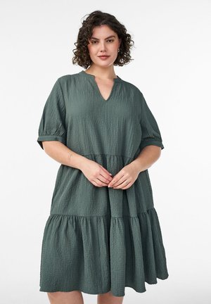 Frau mit lockigem Haar, die ein strukturiertes dunkelgrünes gestuftes Kleid mit Ellbogenlangen Ärmeln trägt und mit locker verschränkten Händen steht.