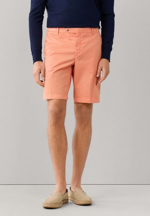 Man die koraalkleurige knielange korte broek, een marineblauw overhemd met lange mouwen en beige instappers draagt, staand tegen een eenvoudige grijze achtergrond.