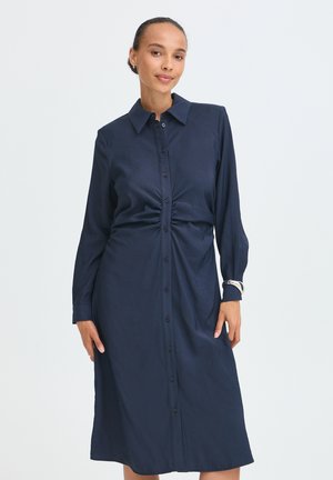 Vrouw met haar naar achter gebonden draagt een marineblauwe midi-jurk met lange mouwen, knopen en geplooide taille, tegen een effen achtergrond.