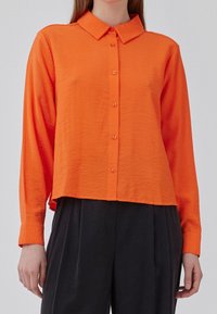 Chemise orange à manches longues avec col, dotée d'un design légèrement raccourci et d'un tissu texturé. Les boutons sont assortis à la couleur de la chemise.