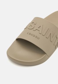Sandale à glissière beige avec une large sangle embossée de "SAINTS LONDON", comprenant une semelle intérieure texturée et un matériau lisse semblable au caoutchouc.