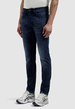 Mann trägt dunkelblaue Slim-Fit-Jeans, weiße Sneakers und ein weißes ärmelloses Shirt, steht vor hellem Hintergrund.