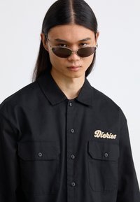 Camicia nera con bottoni, dotata di due tasche frontali, con logo "Dickies" ricamato. Il modello indossa occhiali da sole ovali con montatura argentata.