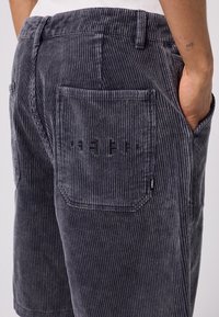 Shorts en velours côtelé gris avec des rayures verticales, dotés d'une poche arrière avec un logo et d'un détail de couture sur le côté, texture douce et design décontracté.