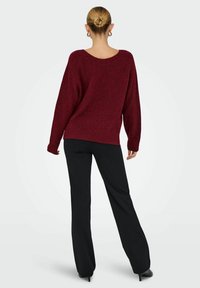 Pull en maille bordeaux avec un col large, des manches longues et une coupe décontractée, associé à un pantalon noir évasé et des talons aiguilles.