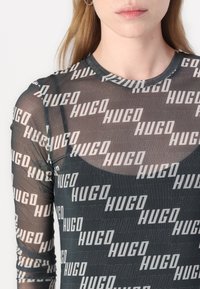 Kerge must pikkade varrukatega särk, millel on korduv "HUGO" logo valgel, ümmargune kaelus, sile tekstuur, kehaülikonda järgiv disain ja minimalismi stiil.