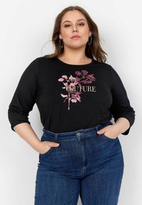 Melna garām piedurknēm kreklā ar rozā ziedu dizainu un vārdu "COUTURE", apvienots ar ziliem augstiem džinsu biksēm.