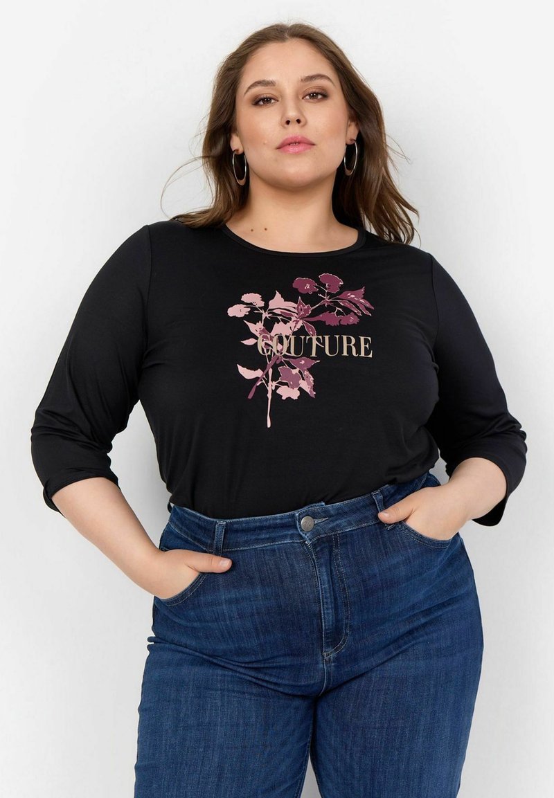 Melna garām piedurknēm kreklā ar rozā ziedu dizainu un vārdu "COUTURE", apvienots ar ziliem augstiem džinsu biksēm.