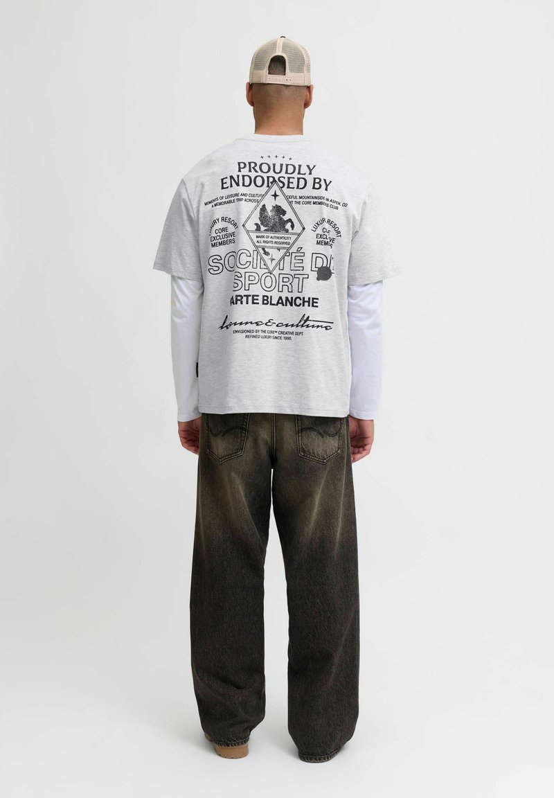 Muž s béžovou čiapkou, bielym tričkom s dlhým rukávom pod šedým tričkom s potlačou a textom, a voľnými tmavými džínami, stojaci na bielom pozadí.