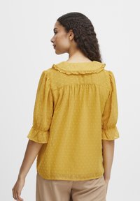 Kollane blouse volangkleidi ja lendlevate varrukatega. Valmistatud kergest, tekstureeritud kangast, millel on väikesed täppide mustrid.