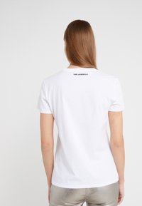 Valkoinen puuvillainen T-paita, jossa on lyhyet hihat ja pyöreä kaula-aukko. Takana, kauluksen lähellä, on musta logo. Sileä pinta.