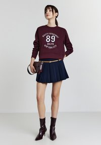 Donna in felpa bordeaux con la scritta "89 BKLYN New York City", gonna a pieghe blu navy con cintura beige, che tiene una borsa bordeaux e indossa stivali con tacco abbinati.