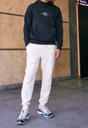 Persona che indossa una felpa con cappuccio nera Calvin Klein, pantaloni jogger bianchi e sneakers bianche e blu, in piedi su un pavimento piastrellato vicino a un muro e a un cartello.