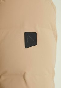 Bežs pufīgs mētelis ar matētu tekstūru, kas aprīkots ar kvadrātveida melnu etiķeti ar logotipu. Audums izskatās gluds un viegli atstarojošs.