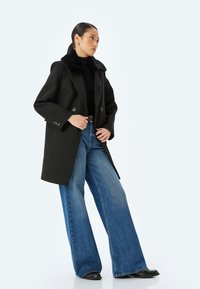 Dobbeltknappet sort frakke med en faux fur krave, brede klapper og to store knapper. Parret med højtaljede, brede blå jeans.