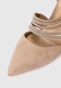 Chaussure en suède beige à bout pointu avec quatre sangles incrustées de strass scintillants sur le dessus.