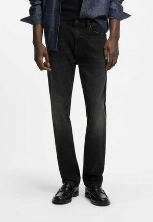 Mann trägt ausgebleichte schwarze Jeans, schwarze Lederloafers und ein dunkelblau kariertes Hemd mit hochgekrempelten Ärmeln.