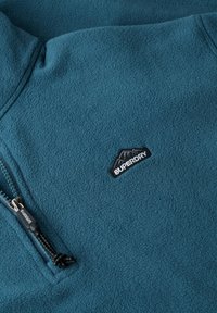 Petrolkleurig fleecejack met een klein zwarte berglogo-patch en "Superdry" tekst op de borst, voorzien van een zwarte rits met trekkoord.