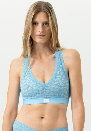 Frau mit langen blonden Haaren, die einen hellblauen Spitzen-Bralette und passende Spitzenunterwäsche vor einem einfarbigen weißen Hintergrund trägt.