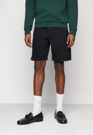 Mann trägt schwarze Shorts, dunkelgrünes Sweatshirt, weiße Socken mit kleinem Logo und schwarze Loafers, steht vor einem einfarbigen Hintergrund.